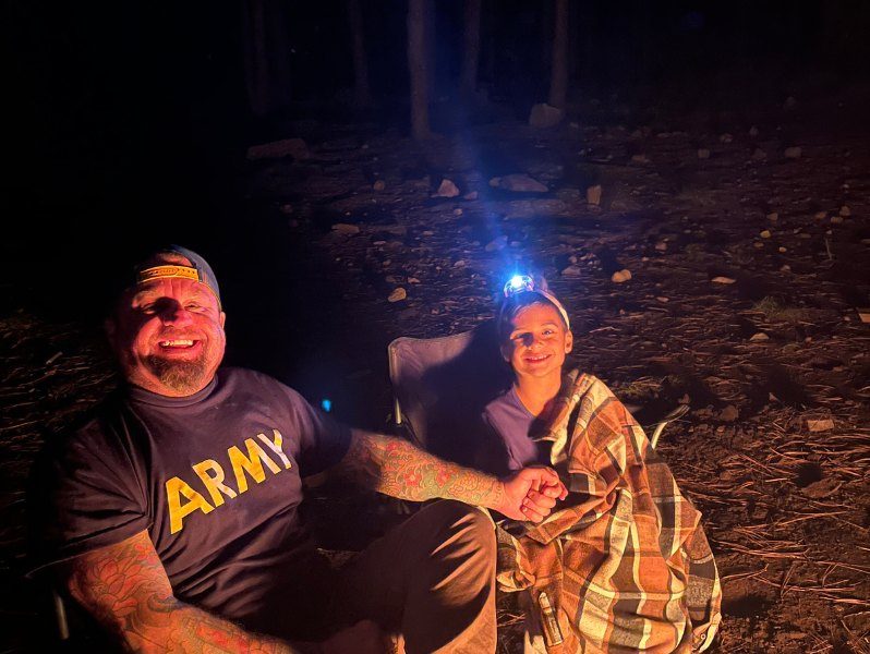 A dad and his son in headlamps laughing by the campfire at night