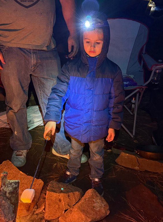 A young boy in a headlamp roasting a marshmallow by the fire