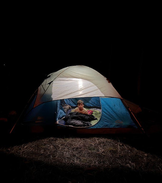 A boy in a lit tent at night, tucked into his sleeping bag